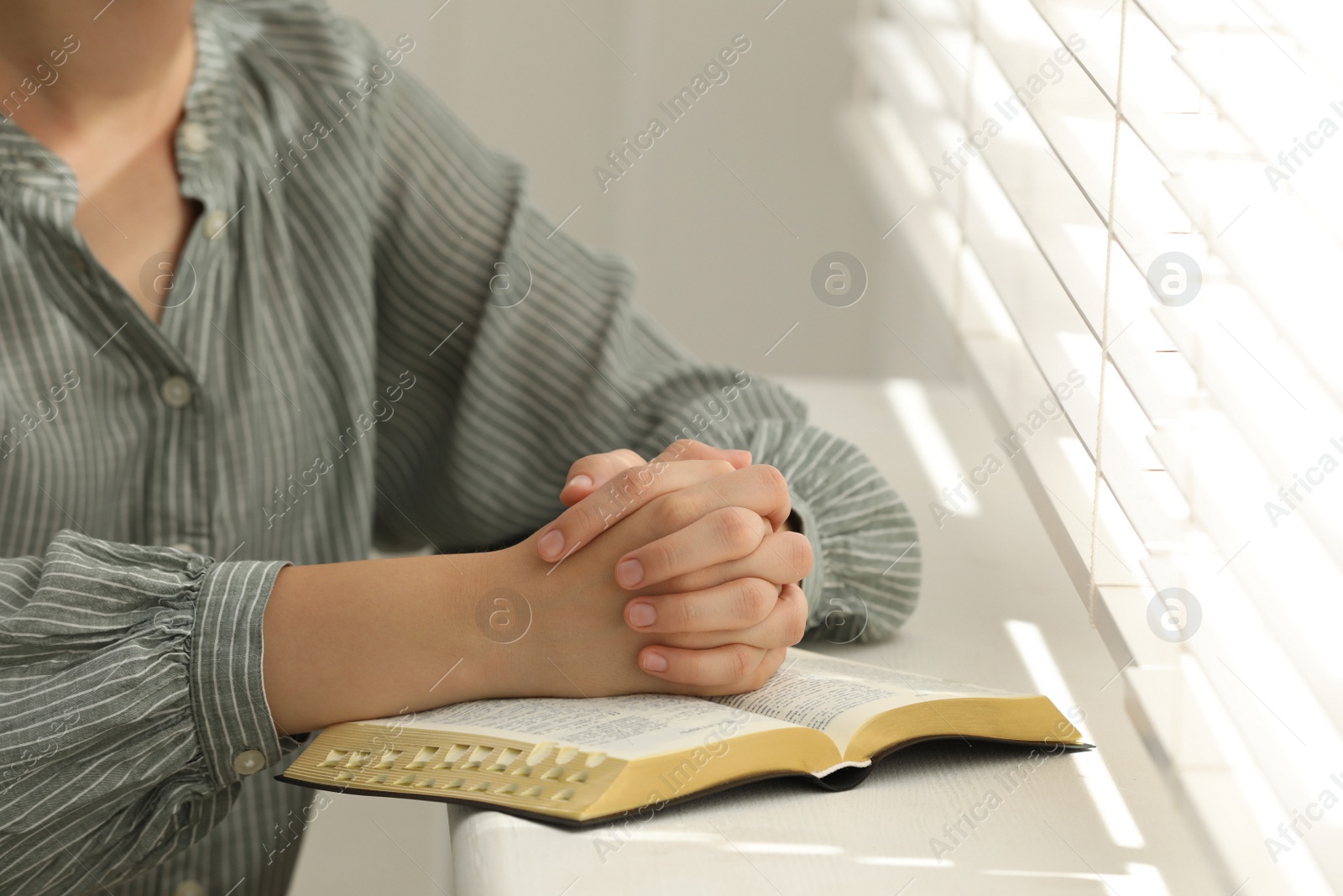 Photo of Religious woman praying over Bible near window indoors, closeup