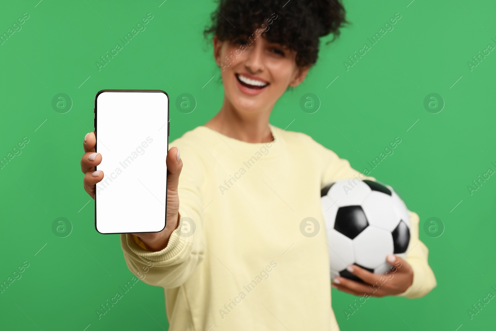 Photo of Happy fan holding football ball and showing smartphone on green background, selective focus