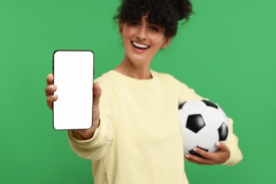 Photo of Happy fan holding football ball and showing smartphone on green background, selective focus