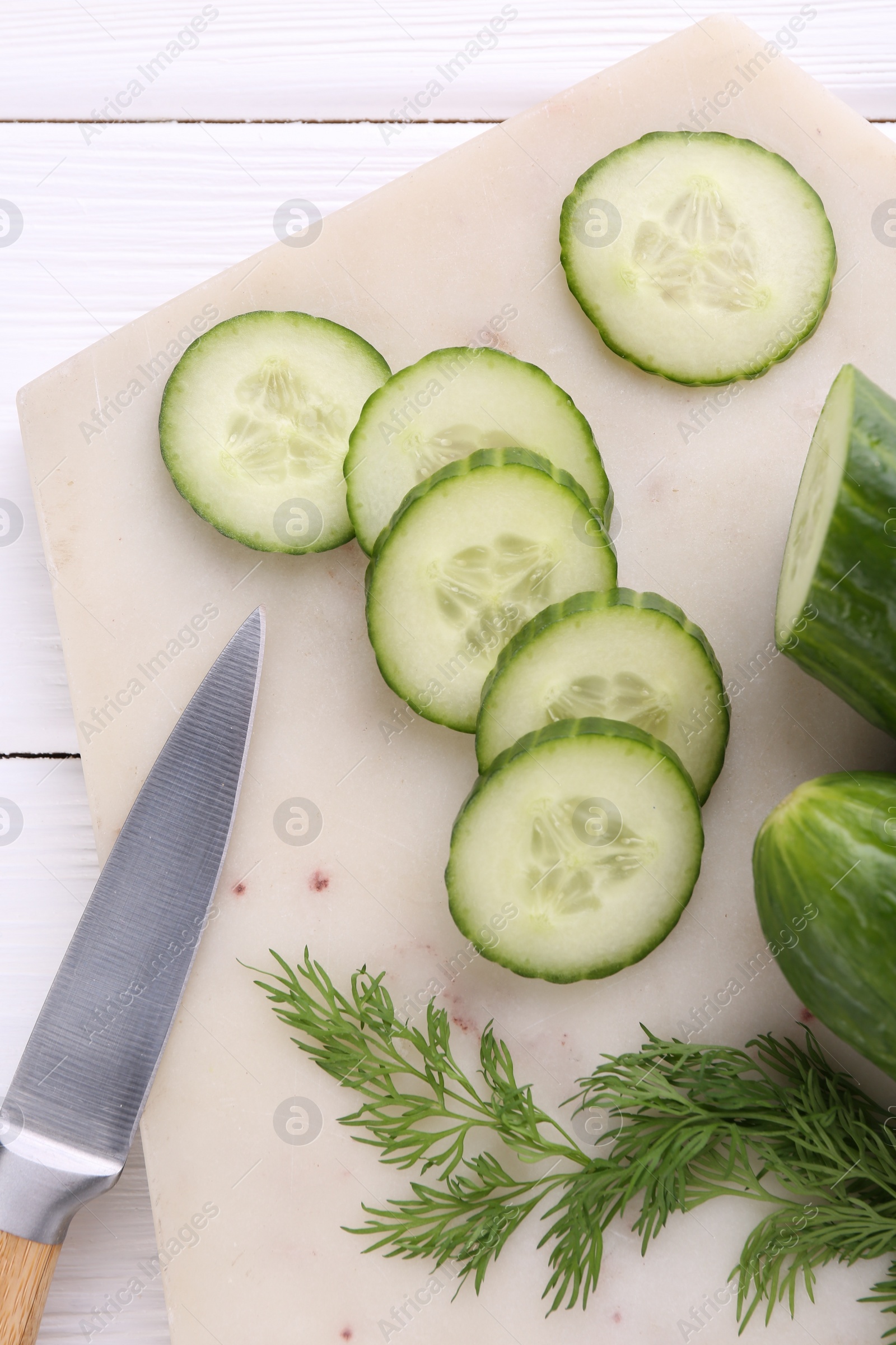 Photo of Cucumbers, dill, knife and marble cutting board on white wooden table, top view