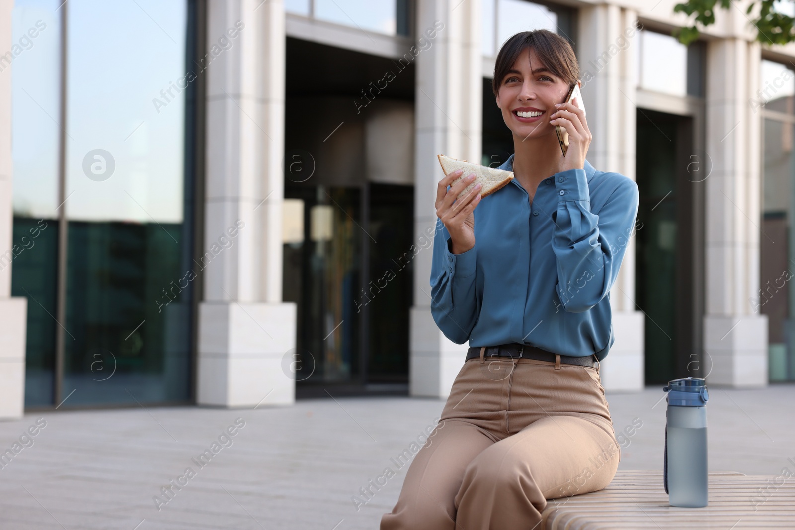 Photo of Happy businesswoman with sandwich talking on smartphone during lunch on bench outdoors