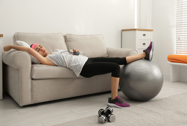 Lazy young woman with sport equipment on sofa at home