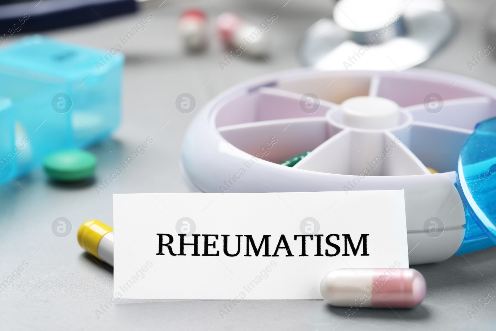 Photo of Sheet of paper with word Rheumatism near container and many different pills on light grey table, closeup