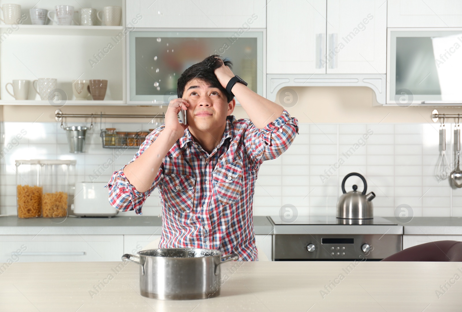 Photo of Emotional man calling plumber near table with saucepan under leaking water from ceiling in kitchen