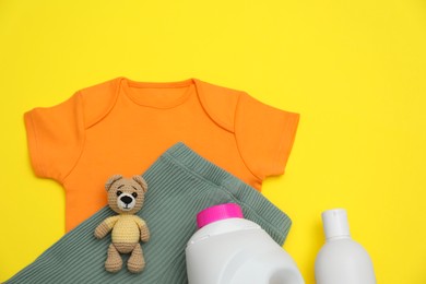 Bottles of laundry detergents, baby clothes and toy bear on yellow background, flat lay