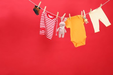 Different baby clothes and toys drying on laundry line against red background. Space for text