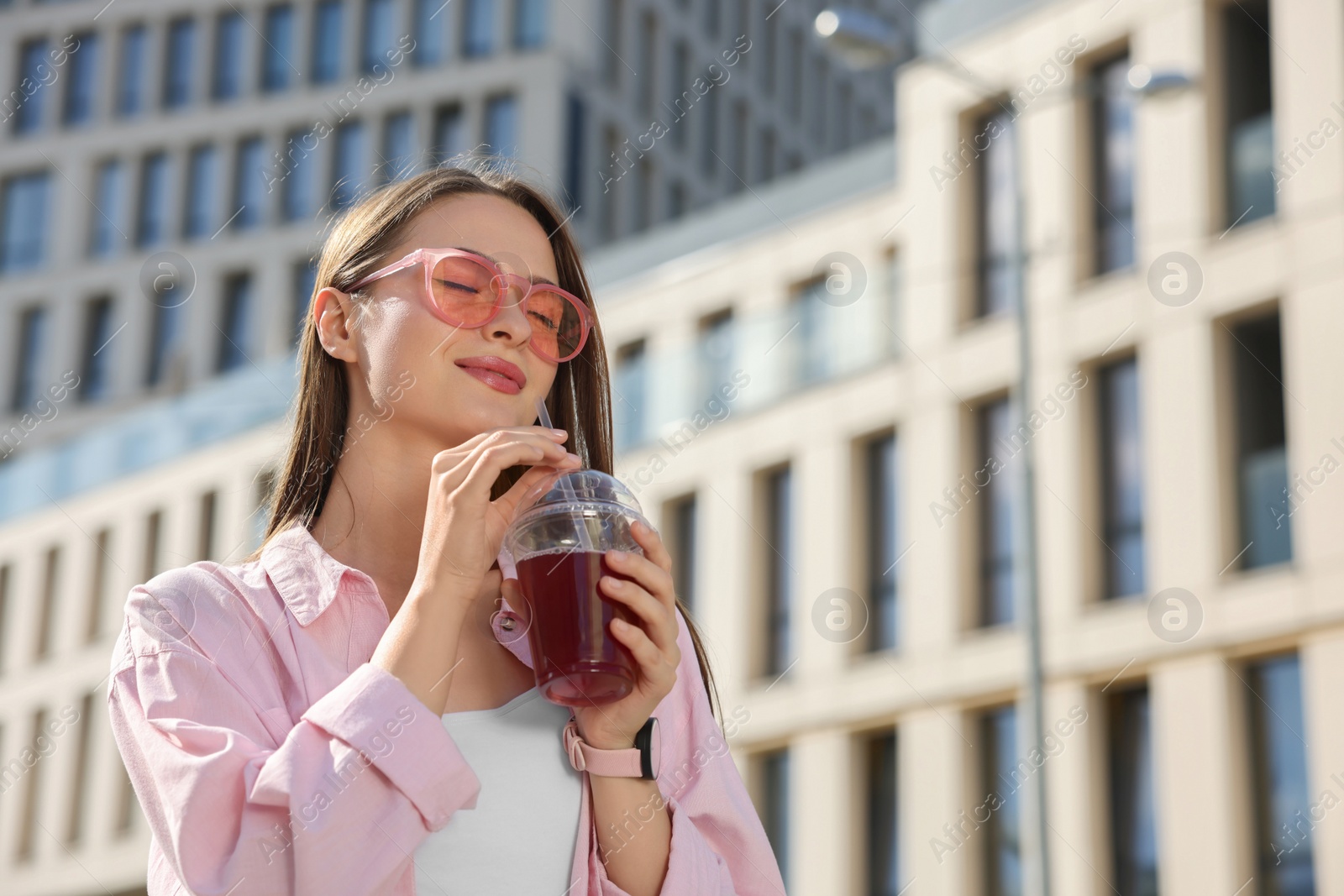 Photo of Young woman in sunglasses with plastic cup of fresh juice outdoors, space for text