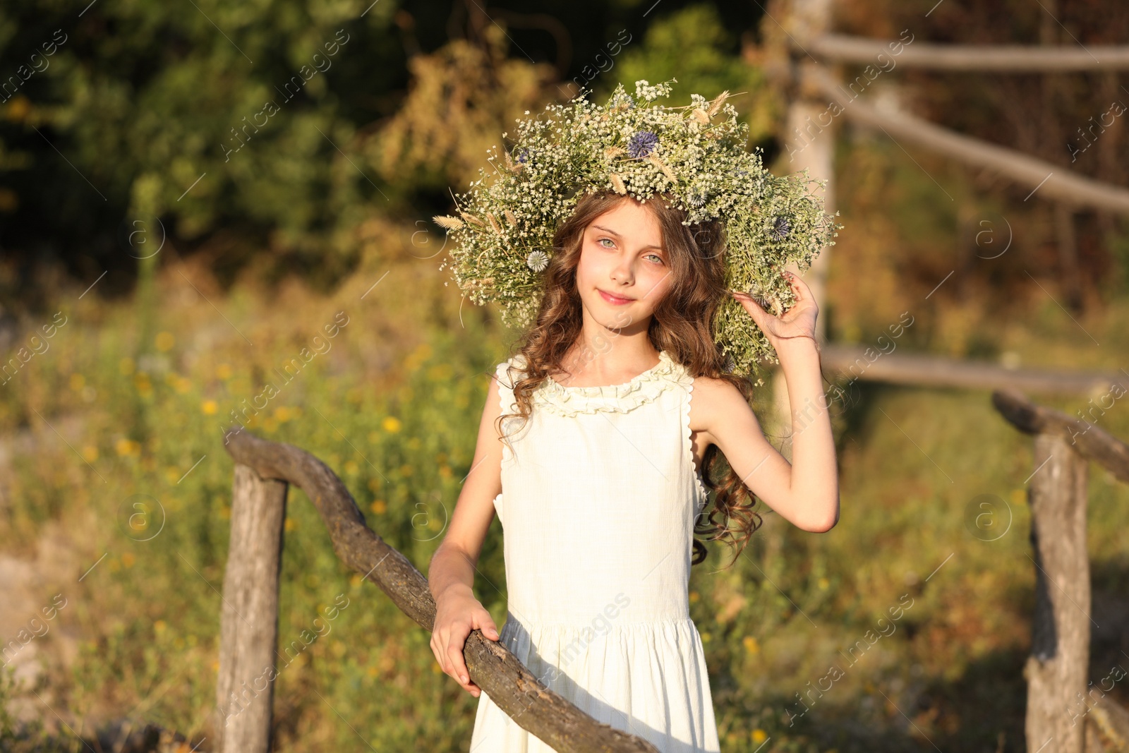 Photo of Cute little girl wearing wreath made of beautiful flowers near wooden fence outdoors on sunny day