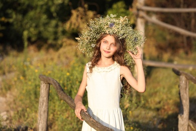 Cute little girl wearing wreath made of beautiful flowers near wooden fence outdoors on sunny day