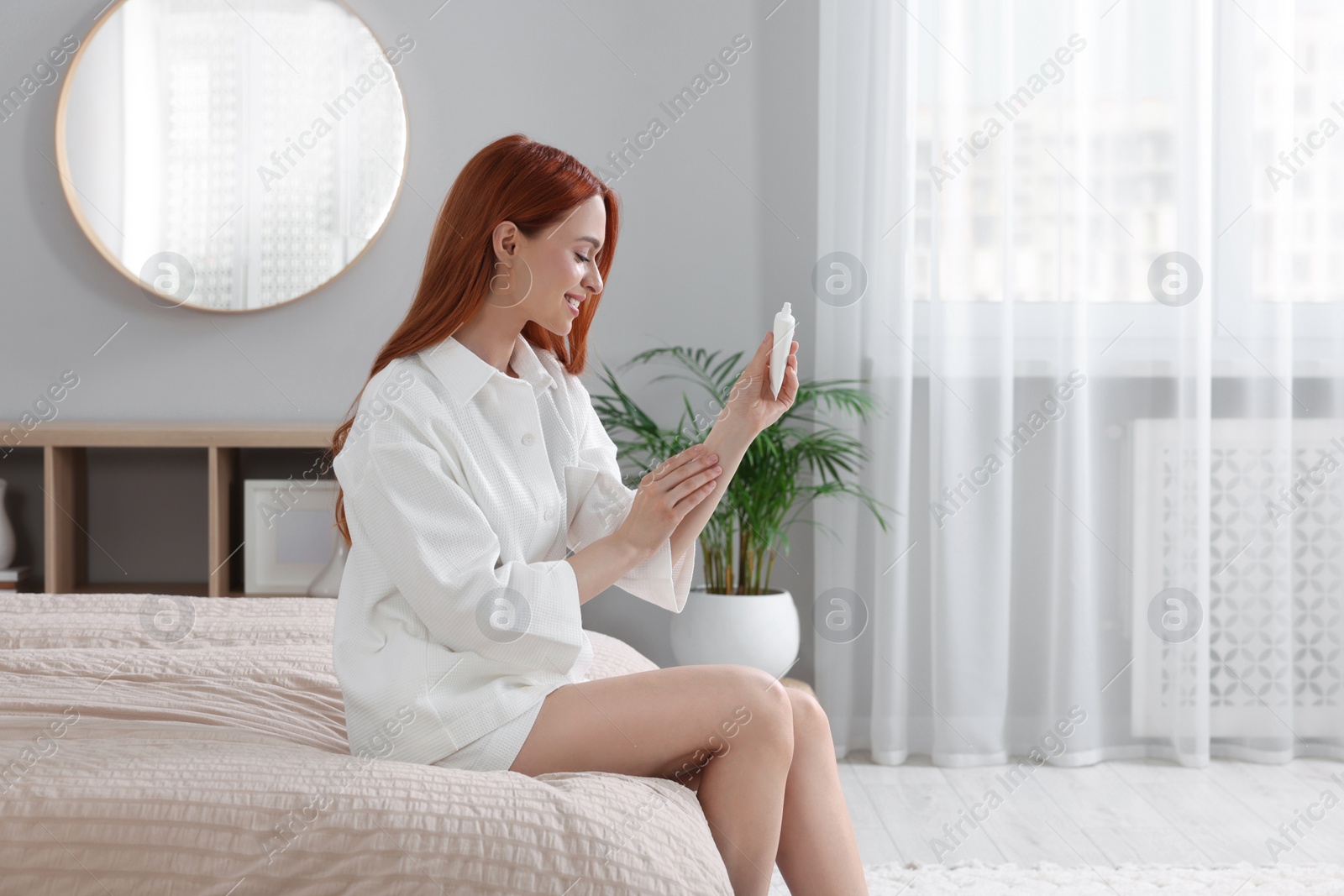 Photo of Beautiful young woman applying body cream onto arms in bedroom, space for text