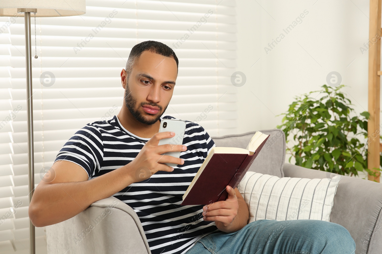 Photo of Young man using smartphone while reading book at home. Internet addiction