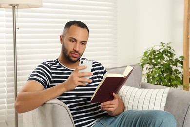 Photo of Young man using smartphone while reading book at home. Internet addiction