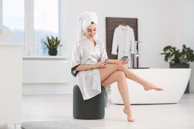 Photo of Beautiful happy woman in stylish bathrobe applying cream on leg in bathroom