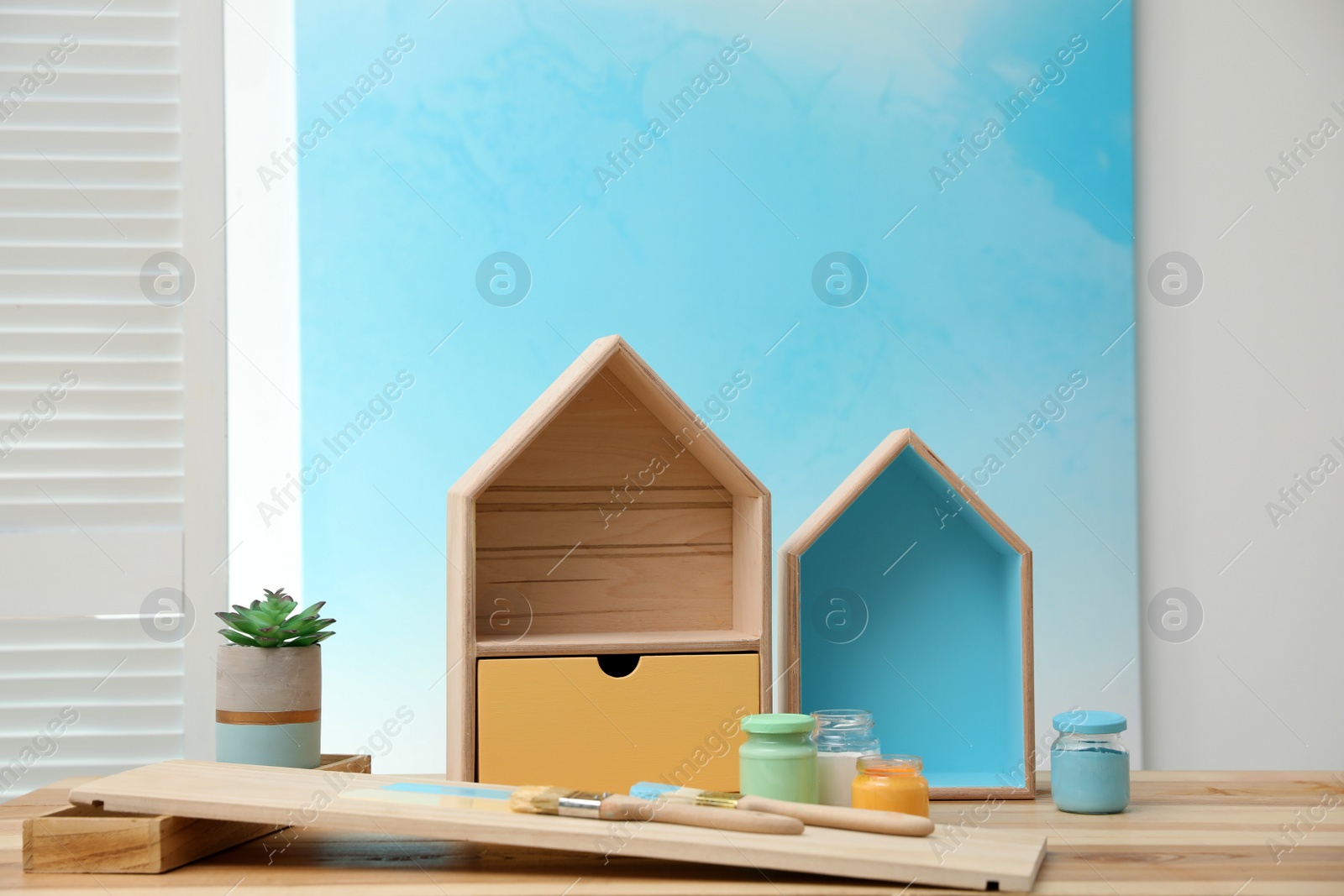 Photo of House shaped shelves, brushes and jars of paints on table indoors