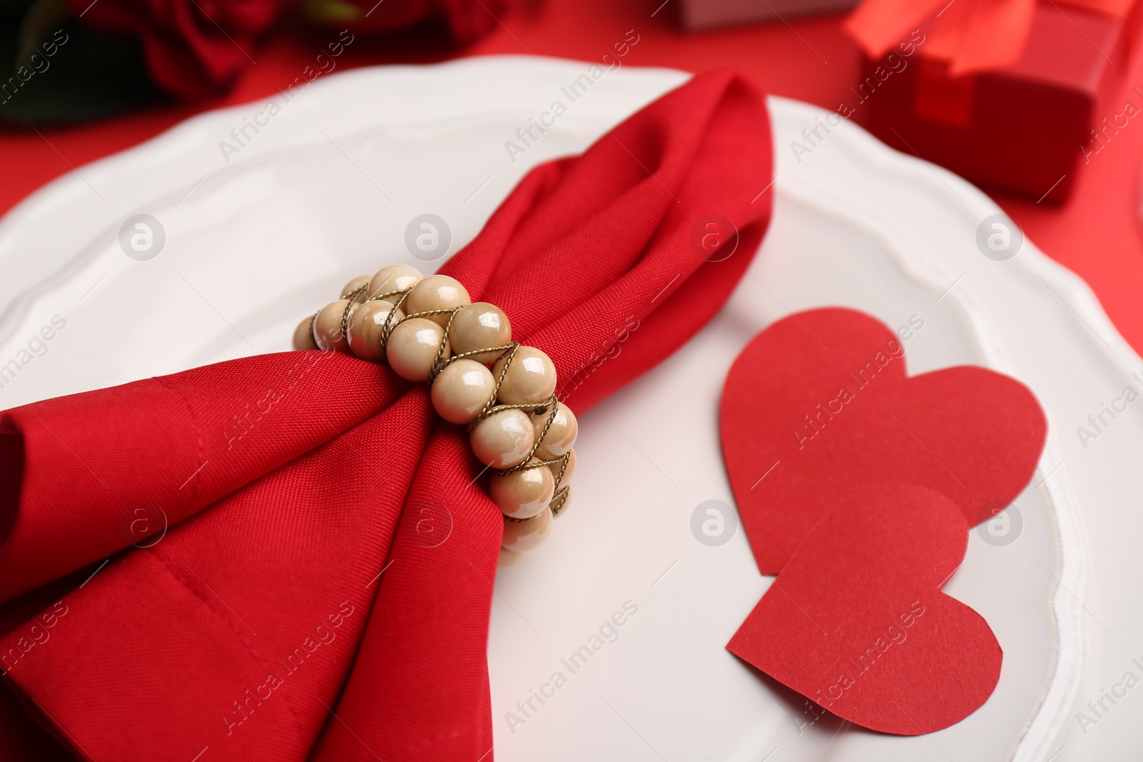 Photo of Plates with napkin, paper hearts and gift box bouquet of roses on red table, closeup. Romantic dinner place setting