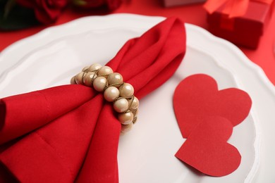 Photo of Plates with napkin, paper hearts and gift box bouquet of roses on red table, closeup. Romantic dinner place setting