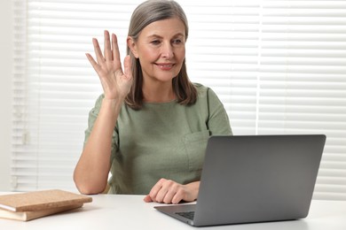 Photo of Happy woman waving hello during video call at table indoors