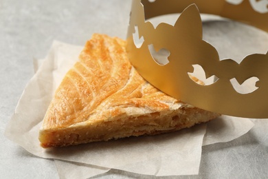 Slice of traditional galette des Rois with paper crown on light grey table, closeup