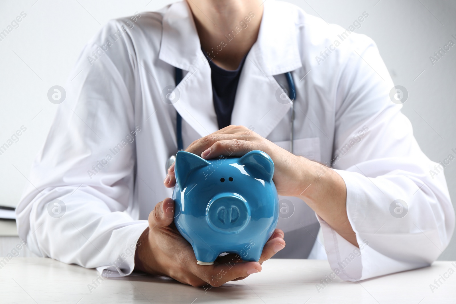 Photo of Doctor with piggybank at white table indoors, closeup. Medical insurance concept