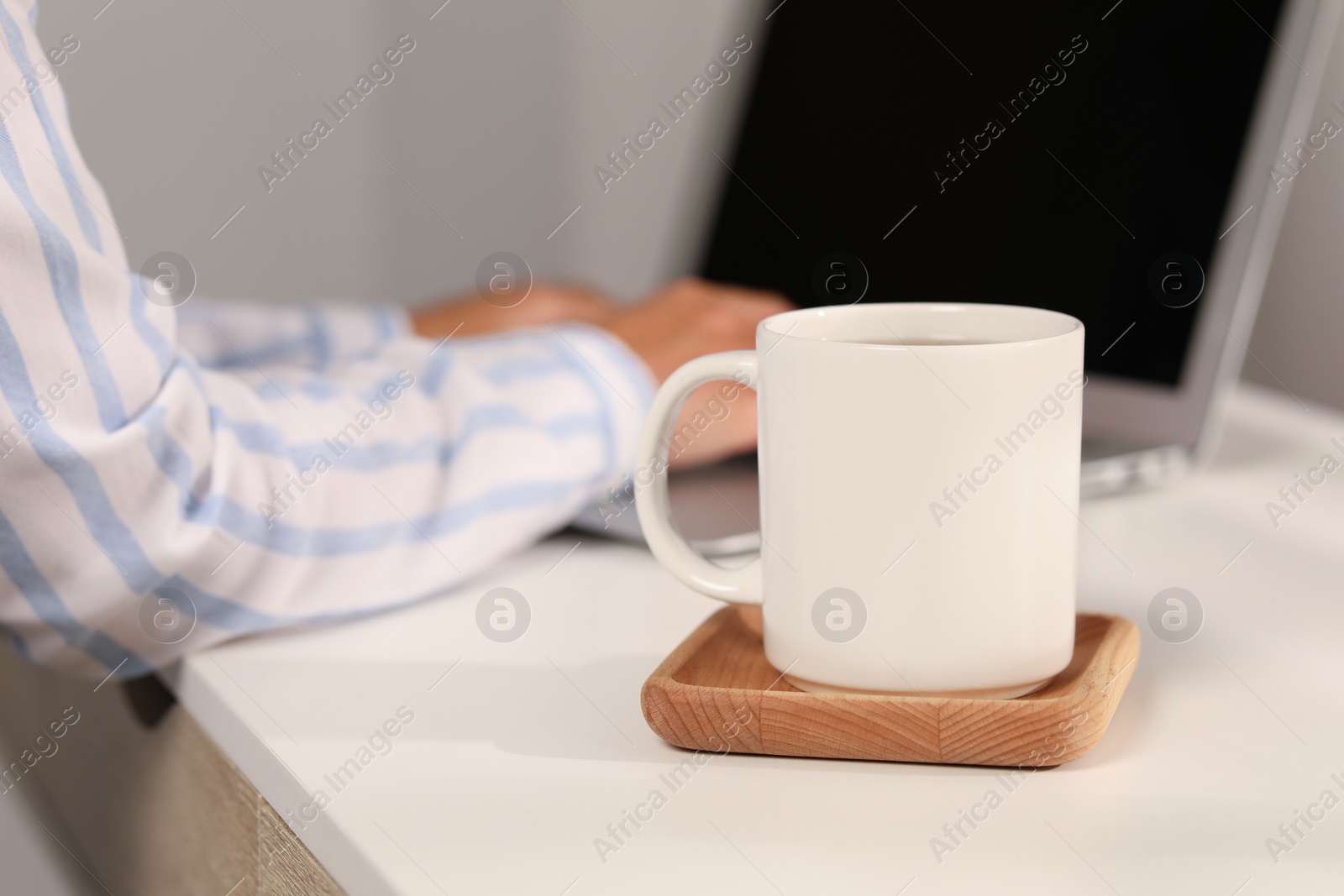 Photo of White ceramic mug on table, selective focus and space for text. Woman working with laptop at workplace, closeup