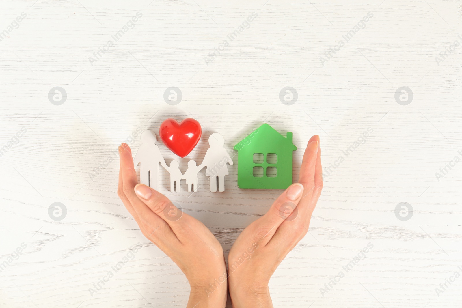 Photo of Woman holding hands near red heart and figures of house and family on white wooden background, top view