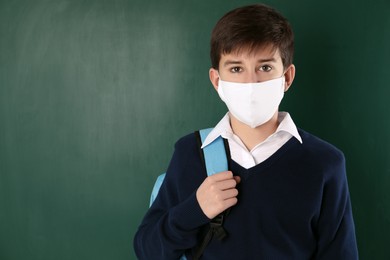 Boy wearing protective mask with backpack near chalkboard, space for text. Child safety