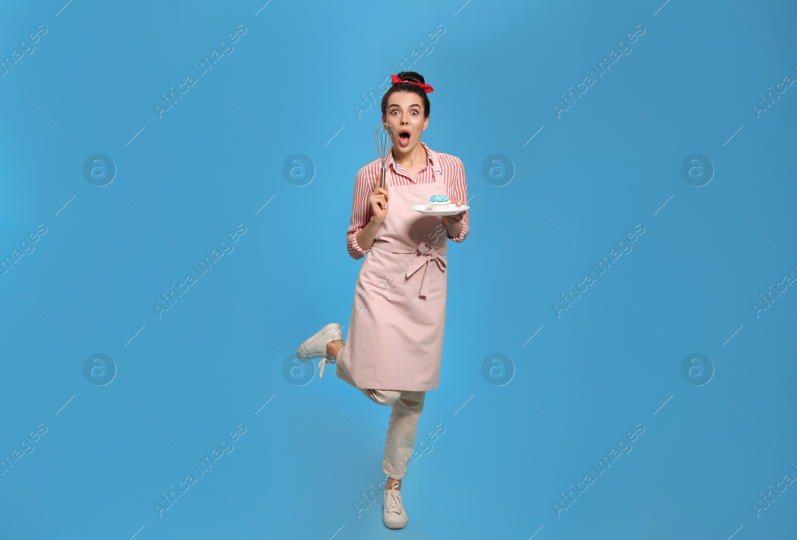 Photo of Emotional housewife with tasty cupcake and whisk on light blue background