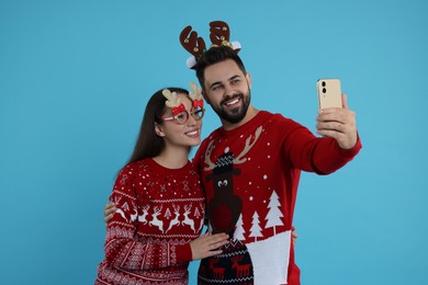 Happy young couple in Christmas sweaters, reindeer headband and party glasses taking selfie on light blue background