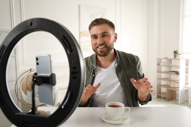 Photo of Blogger with cup of tea recording video at table indoors. Using ring lamp and smartphone