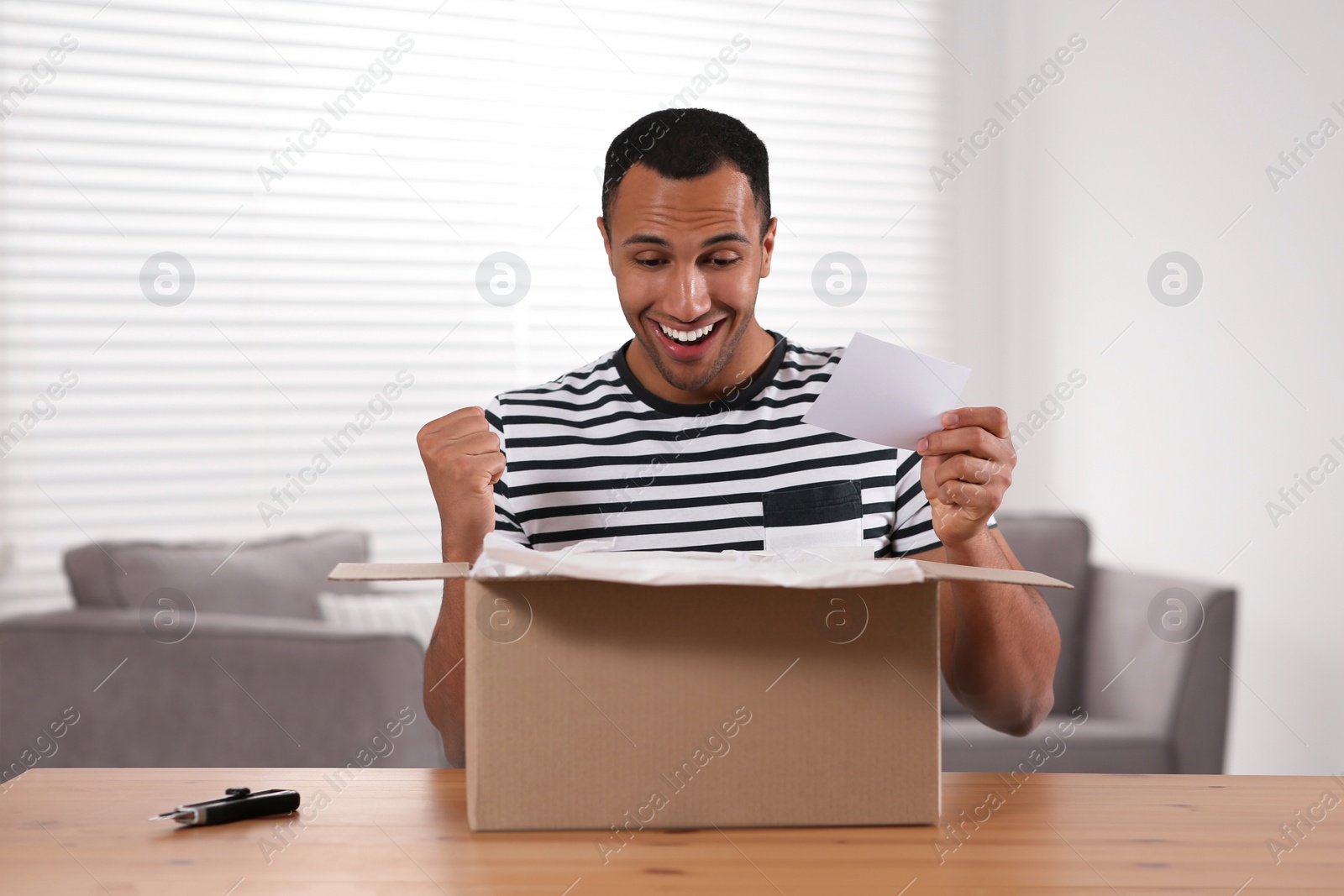 Photo of Emotional young man with greeting card near parcel at table indoors. Internet shopping