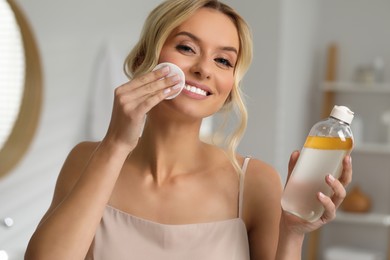 Photo of Smiling woman removing makeup with cotton pad and holding bottle indoors