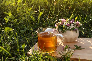 Cup of aromatic herbal tea and ceramic mortar with different wildflowers on green grass outdoors