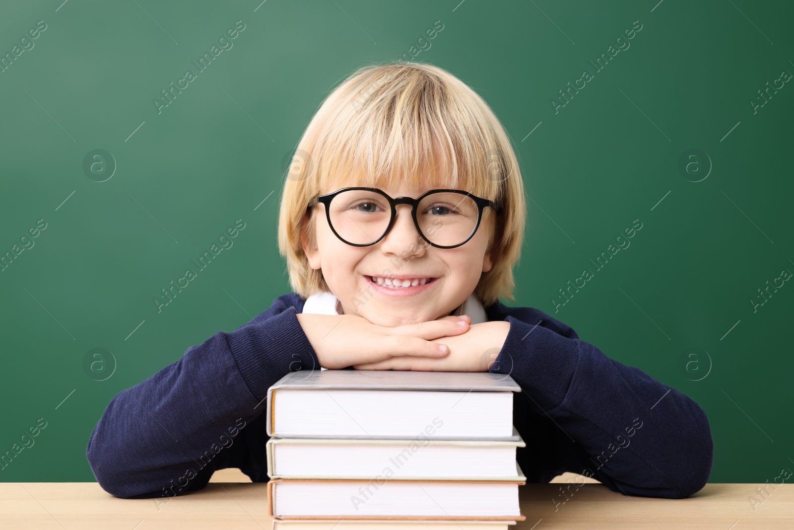 Photo of Happy little school child sitting at desk with books near chalkboard