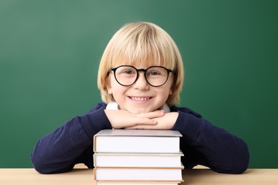 Happy little school child sitting at desk with books near chalkboard