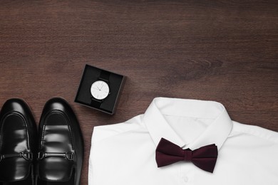 Photo of White shirt with stylish burgundy bow tie, wristwatch and shoes on wooden background, flat lay. Space for text