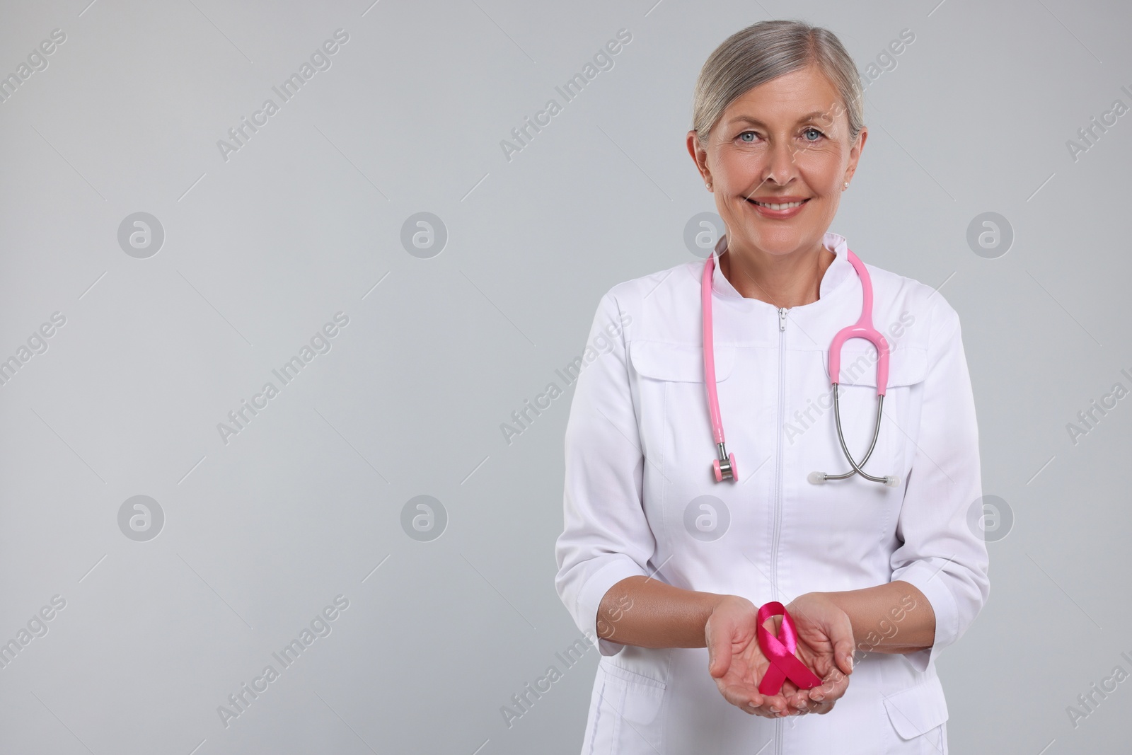 Photo of Doctor with stethoscope holding pink ribbon on light grey background, space for text. Breast cancer awareness