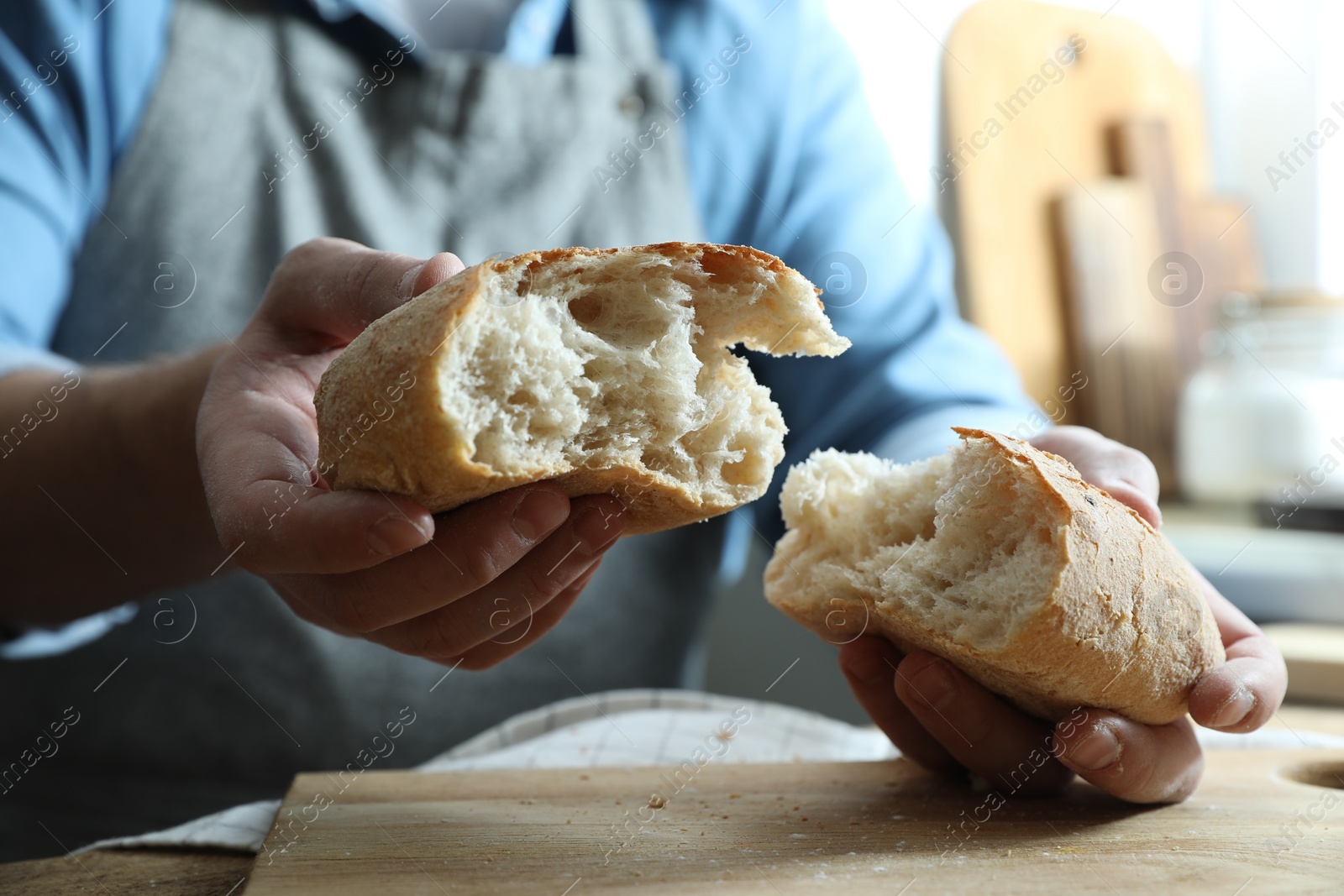 Photo of Man breaking loaf of fresh bread at wooden table indoors, closeup