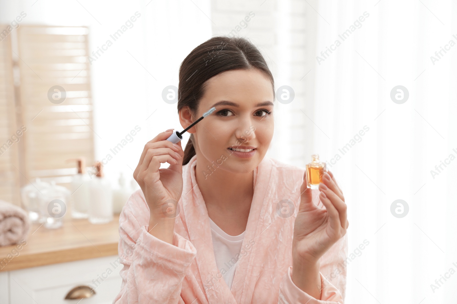 Photo of Beautiful woman applying oil onto her eyelashes indoors