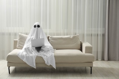 Photo of Creepy ghost. Person covered with white sheet using laptop on sofa at home, space for text