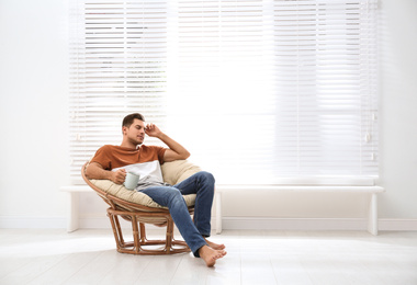 Attractive man relaxing in papasan chair near window at home. Space for text