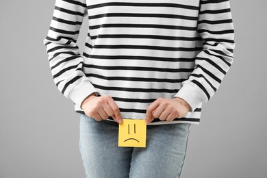 Photo of Cystitis. Woman holding sticky note with drawn sad face on grey background, closeup