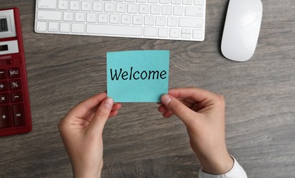 Image of Woman holding paper note with word Welcome over her office desk, top view