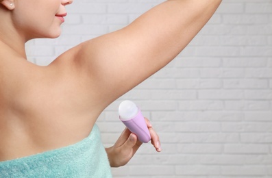 Photo of Young woman applying roll-on deodorant to armpit against brick wall, closeup