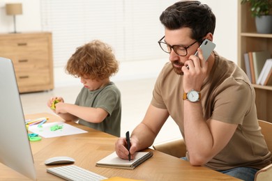 Photo of Man working remotely at home. Busy father talking on smartphone while his son using play dough at desk