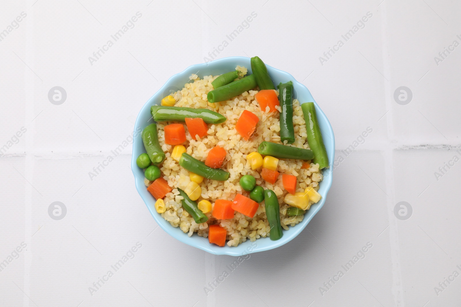 Photo of Delicious bulgur with vegetables in bowl on white tiled table, top view