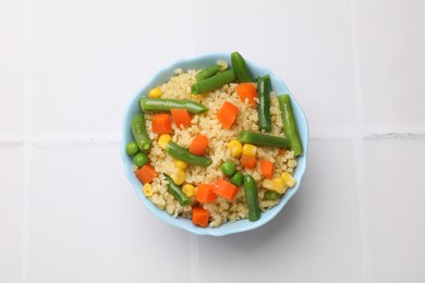 Delicious bulgur with vegetables in bowl on white tiled table, top view