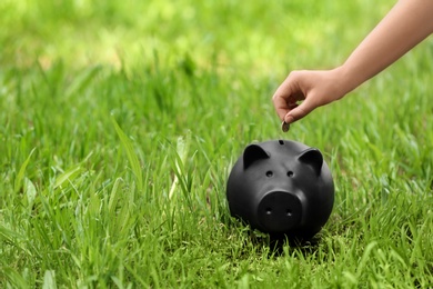 Young woman putting coin into piggy bank on green grass outdoors, closeup. Space for text