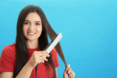 Young woman using hair iron on blue background, space for text