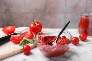 Photo of Glass bowl of tasty tomato paste with spoon and ingredients on white marble table