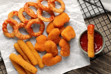 Photo of Tasty chicken nuggets, fried onion rings, cheese sticks and ketchup on wooden table, top view
